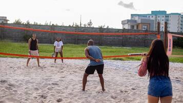 A beach volleyball game with players on a sandy court, a net, and a building in the background.