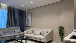 Un salon moderne avec un canapé gris, une table basse, des spots encastrés au plafond, une grande fenêtre avec des rideaux et une plante en pot.