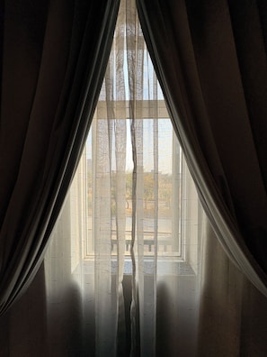 Quarto com janela coberta por cortinas leves, piso de madeira e vista para árvores e um edifício lá fora.