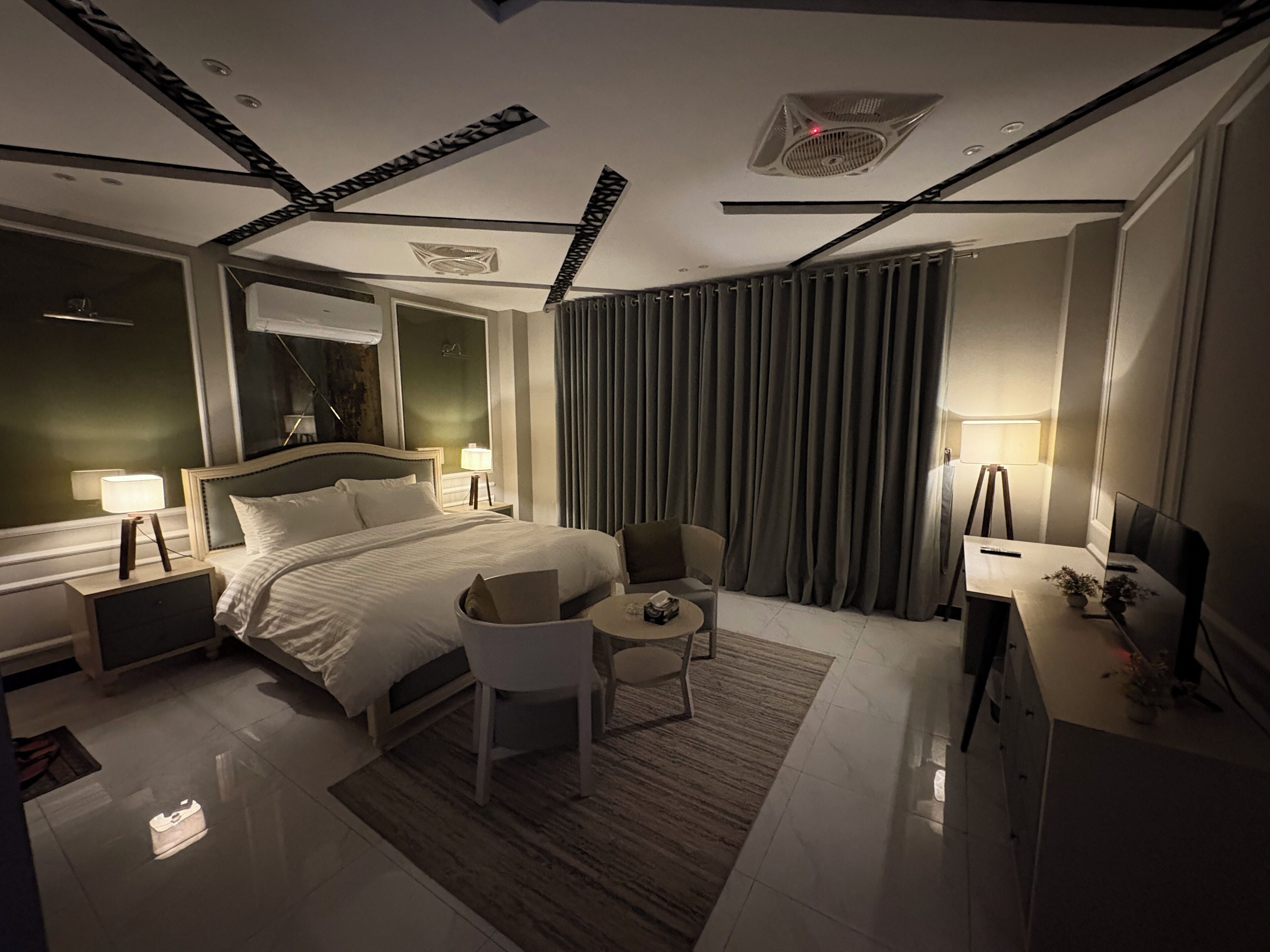 A modern hotel room with a large bed, bedside tables, a desk, a chair, a small table, and a TV.