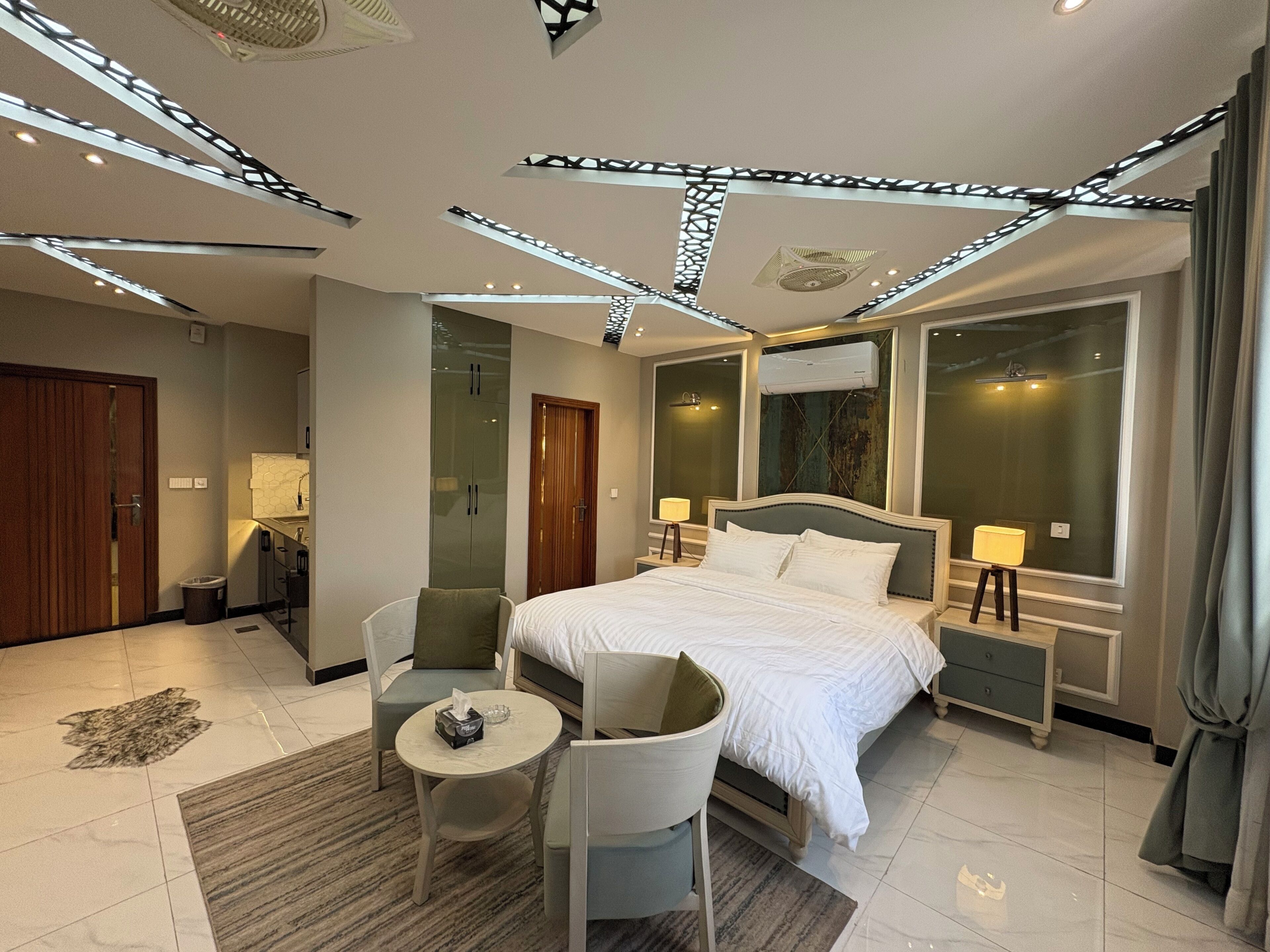A modern hotel room with a large bed, two bedside tables, a chair, a small round table, and a large window.