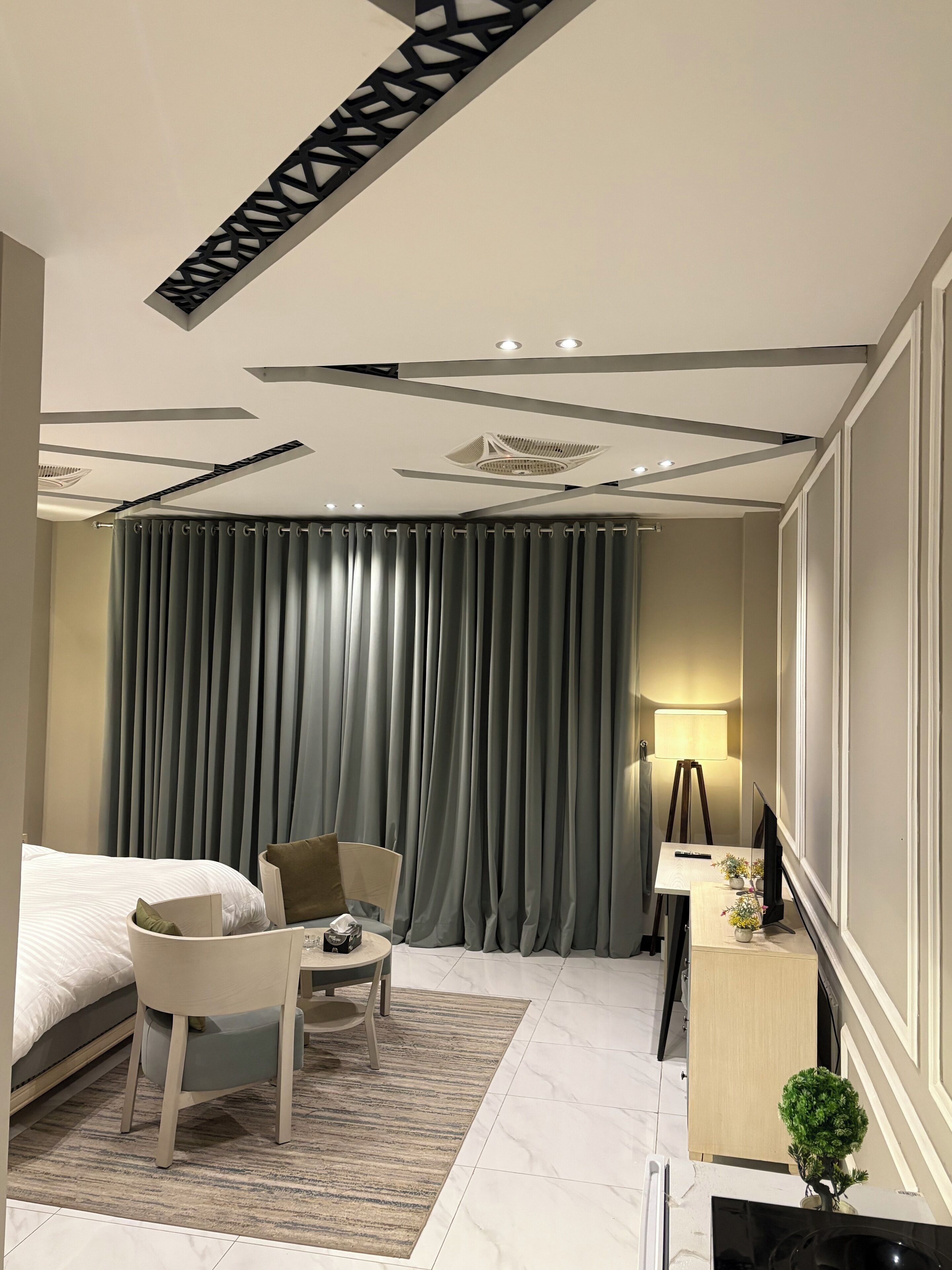 Una habitación de hotel moderna con cama, escritorio con lámpara, silla y una mesita con control remoto y flores.