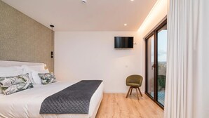 Un dormitorio con cama, televisor montado en la pared, una silla y puerta corrediza de vidrio que da a un balcón.