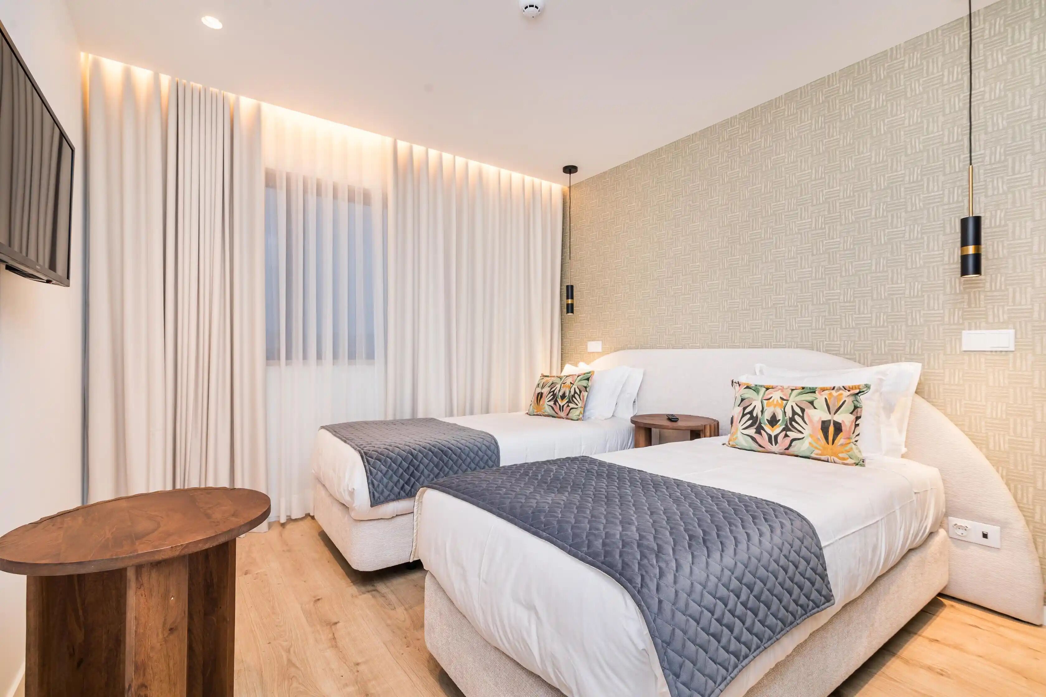 Moderni hotellihuone, jossa on kaksi sänkyä, puinen yöpöytä, televisio ja kuvioitu seinä.