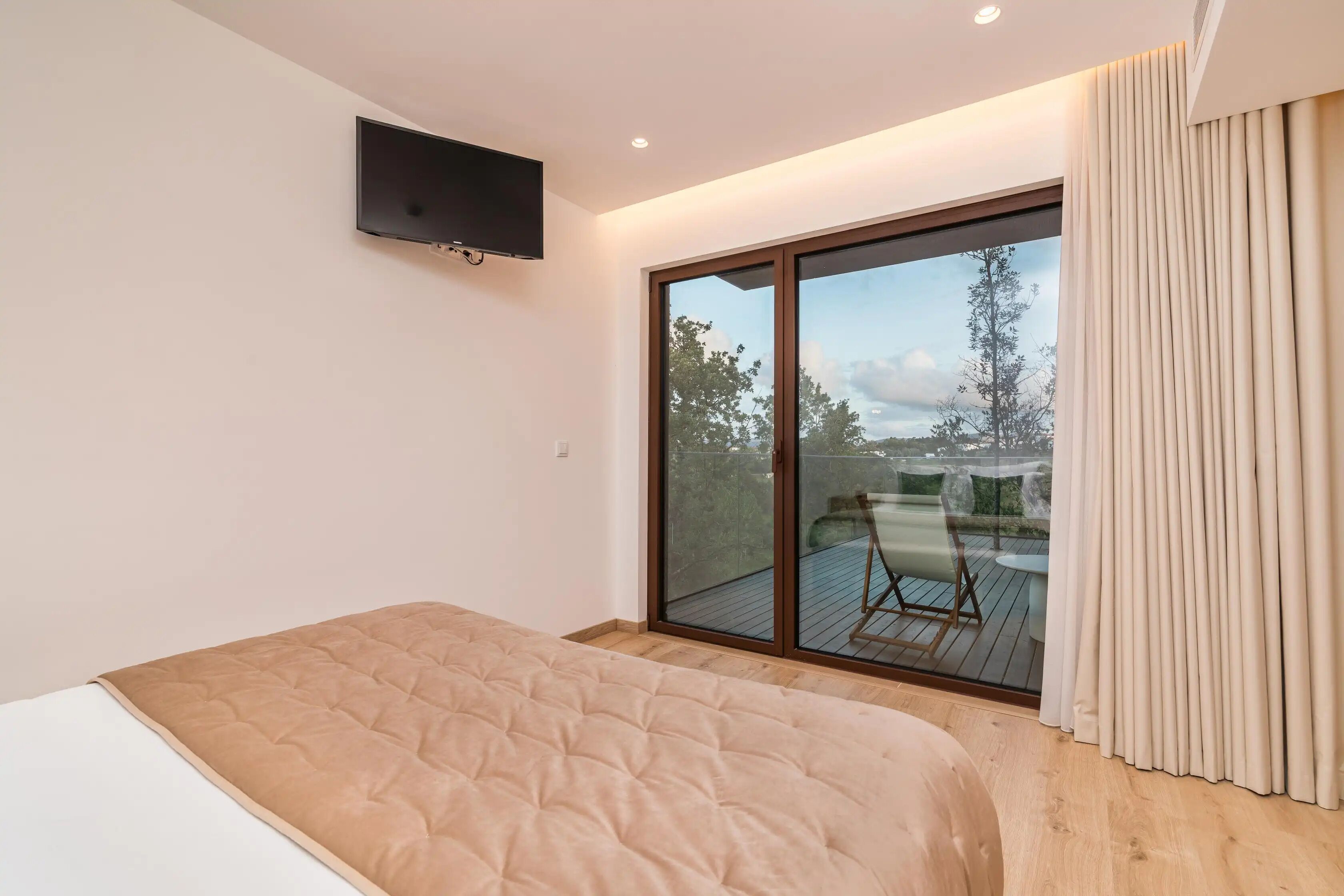 A bedroom with a bed, a TV mounted on the wall, and a sliding glass door leading to an outdoor deck.