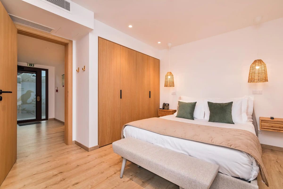 A bedroom with a bed, bench, wardrobe, bedside table, lamp, and a door leading to another room.