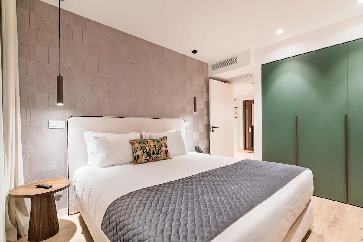A modern hotel room with a large bed, a bedside table, a green wardrobe, and a patterned wall.