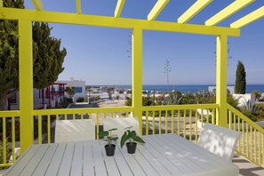 Ein weißer Tisch mit zwei Blumentöpfen, einer gelben Pergola und Blick auf das Meer und Gebäude.
