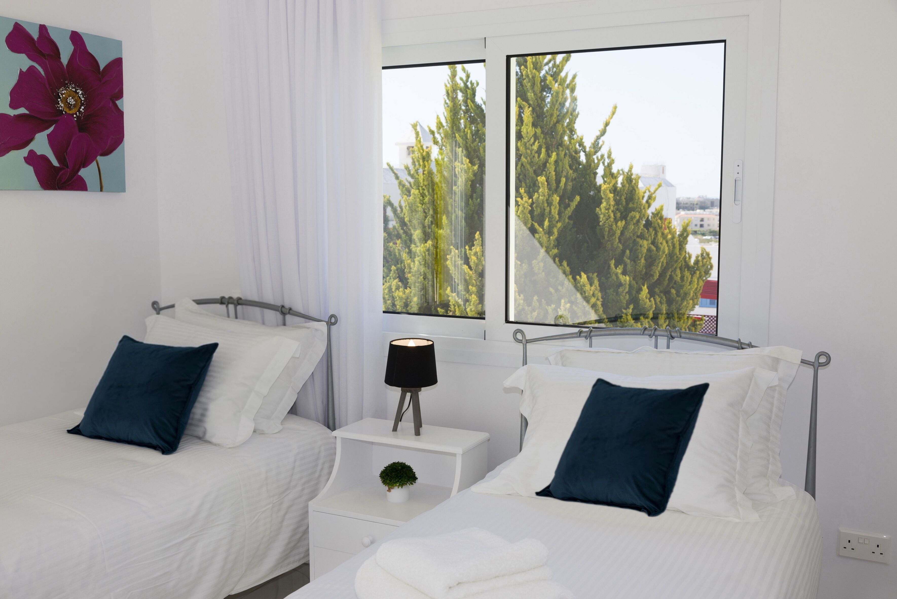Un dormitorio con dos camas, una mesita de noche con lámpara y una ventana con vistas a árboles.