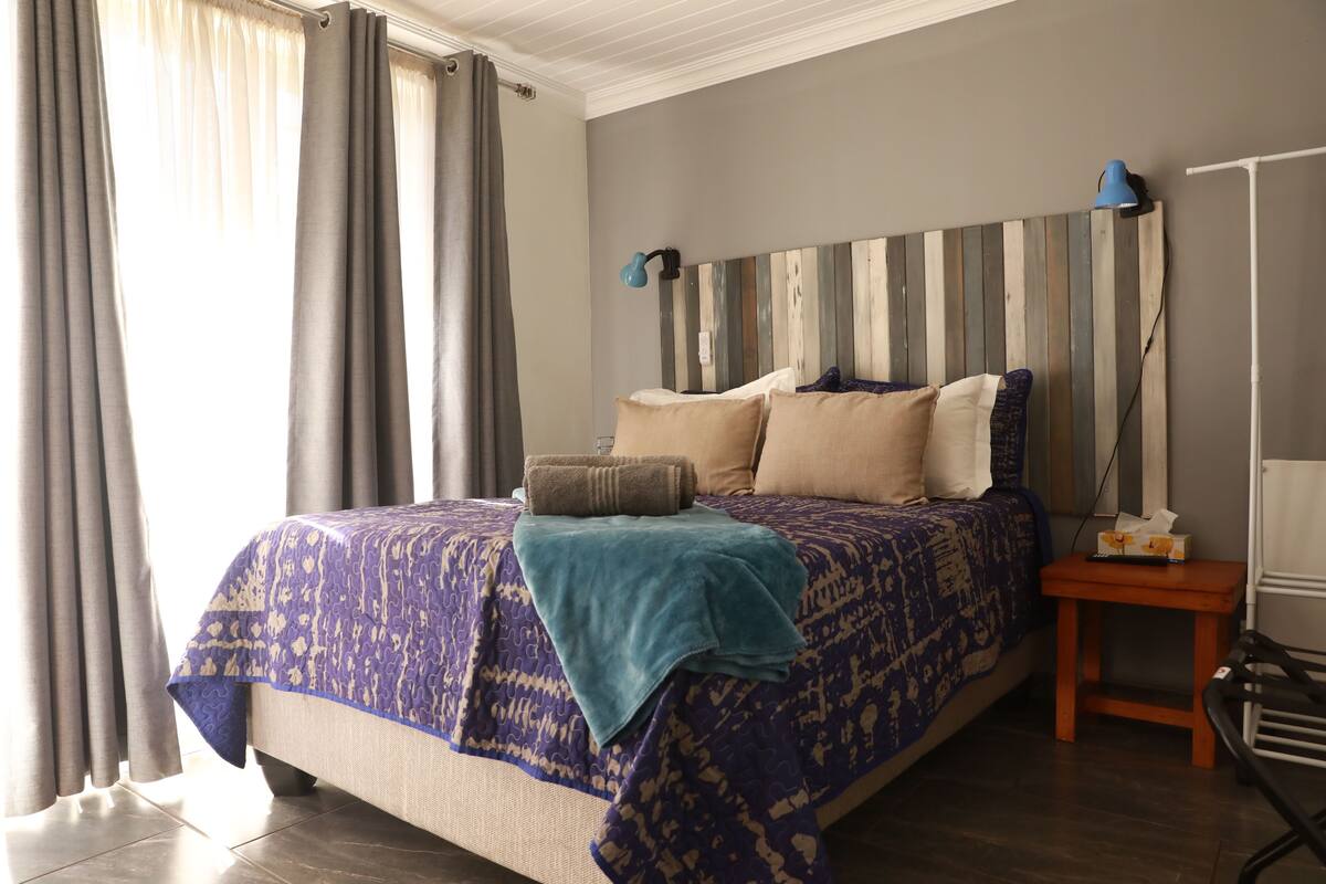 A bedroom with a bed, pillows, a bedside table, a lamp, and curtains.