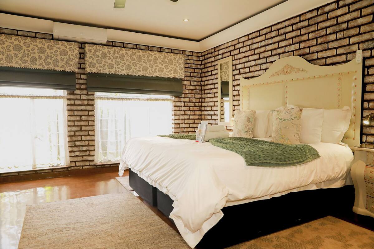 A bedroom with a brick wall, a large bed with white bedding, a nightstand, and a window with patterned curtains.