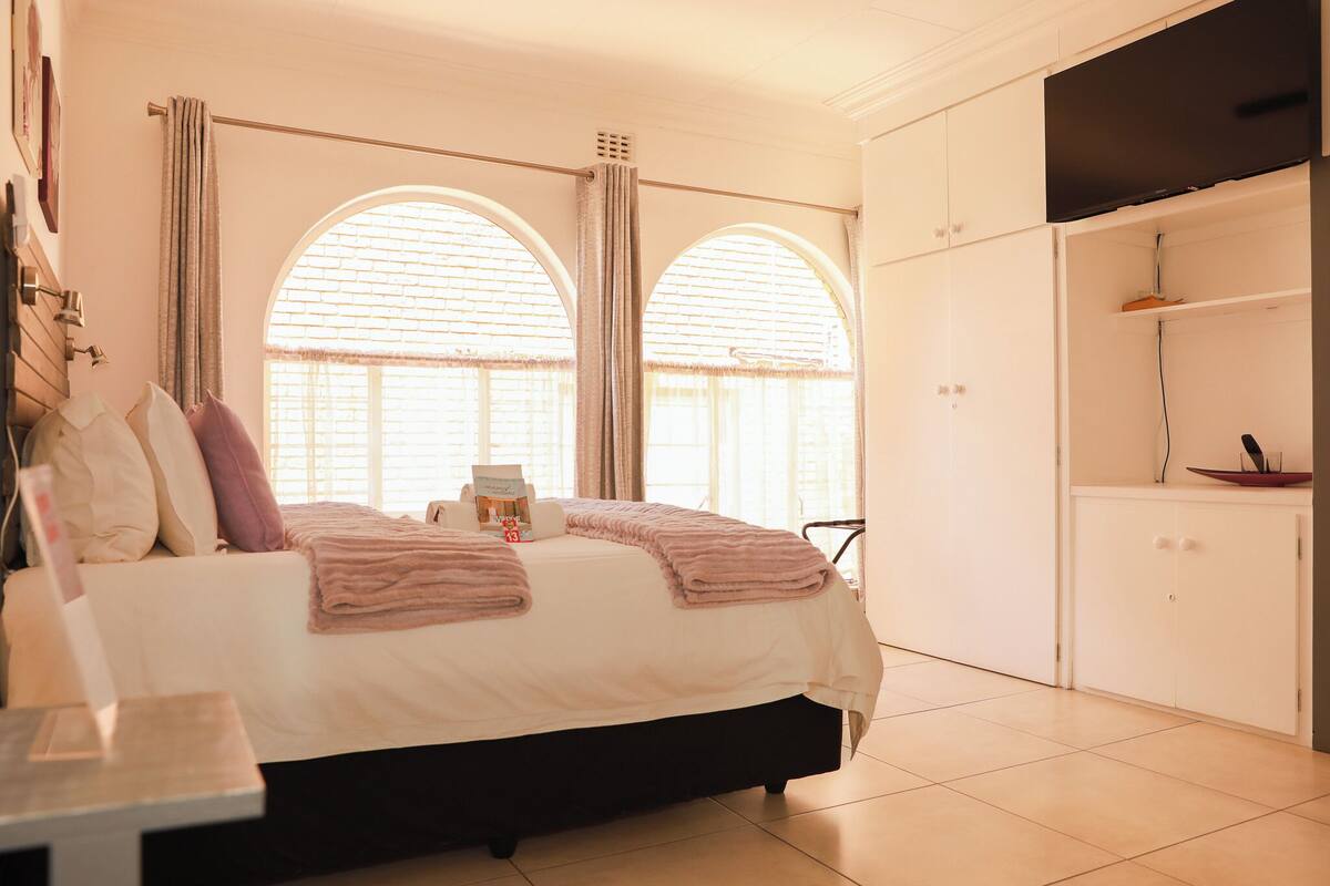 A bedroom with a bed, two windows with curtains, a bedside table, a wardrobe, and a television.