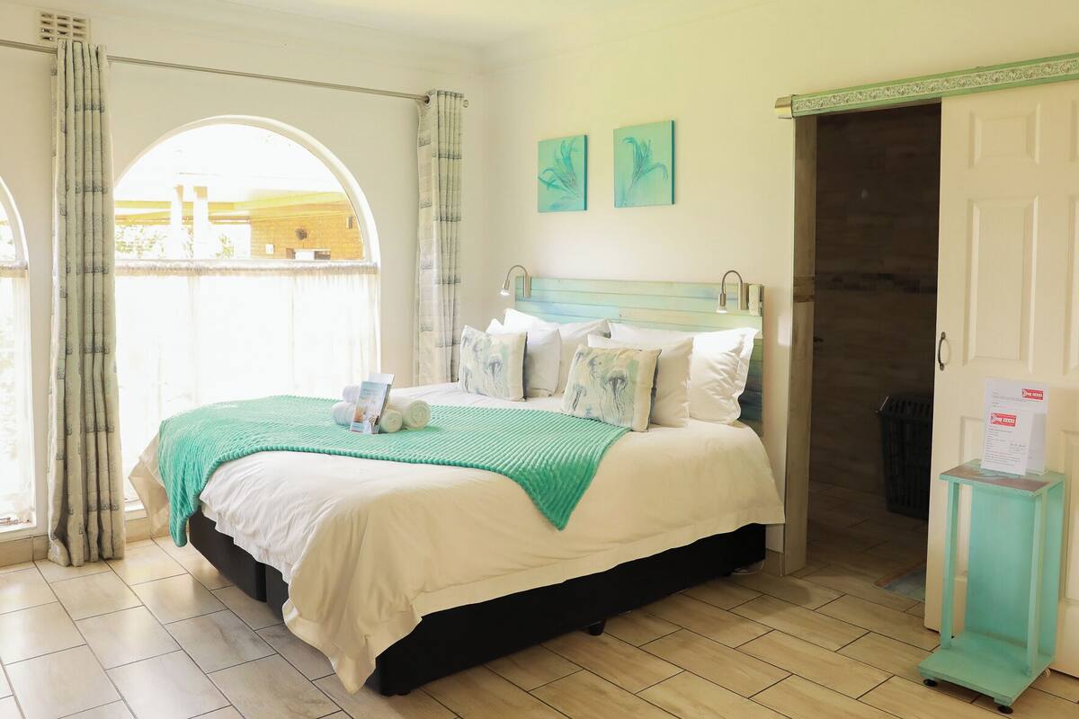 A hotel room with a bed, a window with curtains, a door, and a small table with a sign.