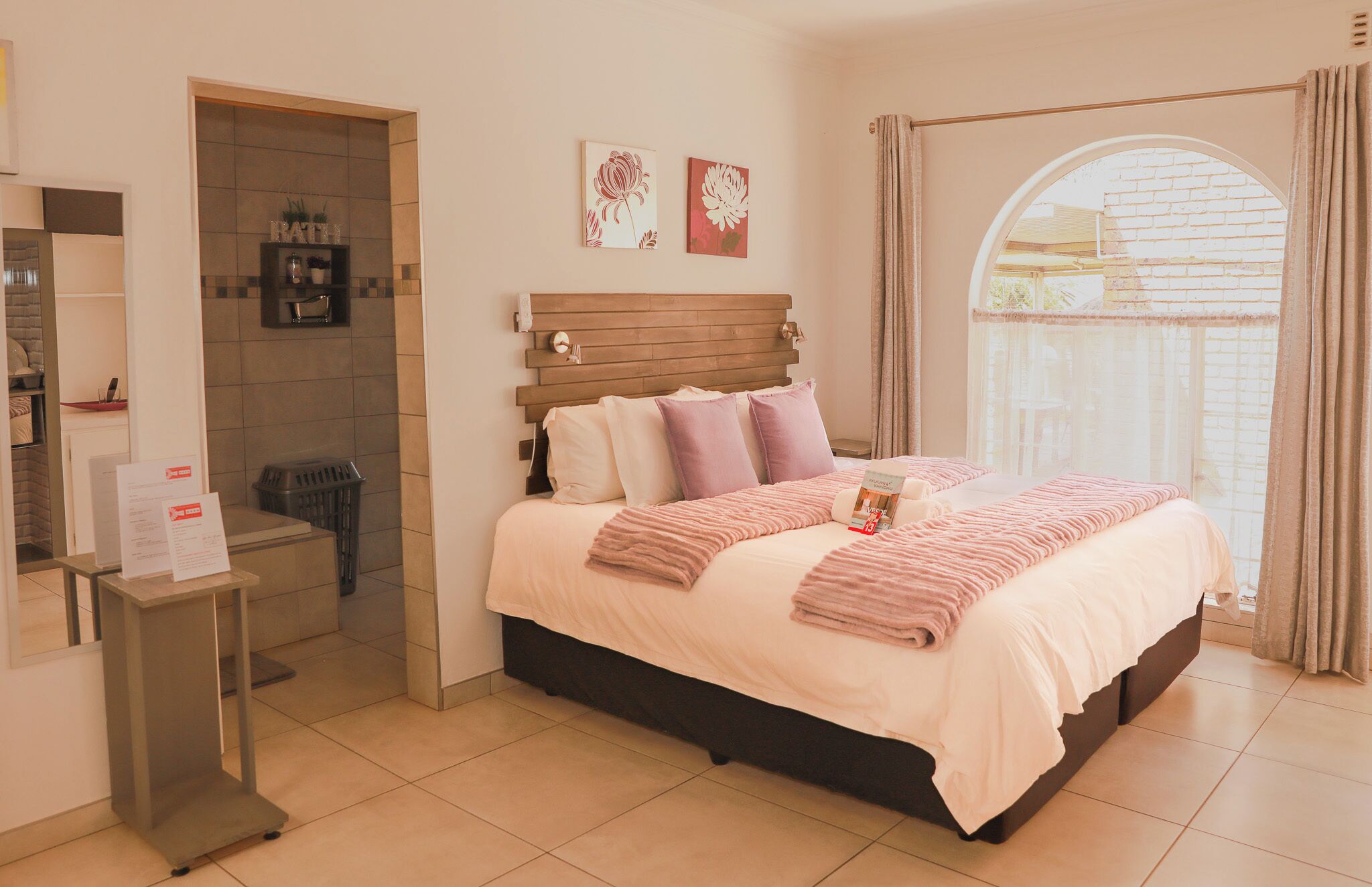 A hotel room with a bed, a small table, a mirror, a bathroom, and a view through a window.
