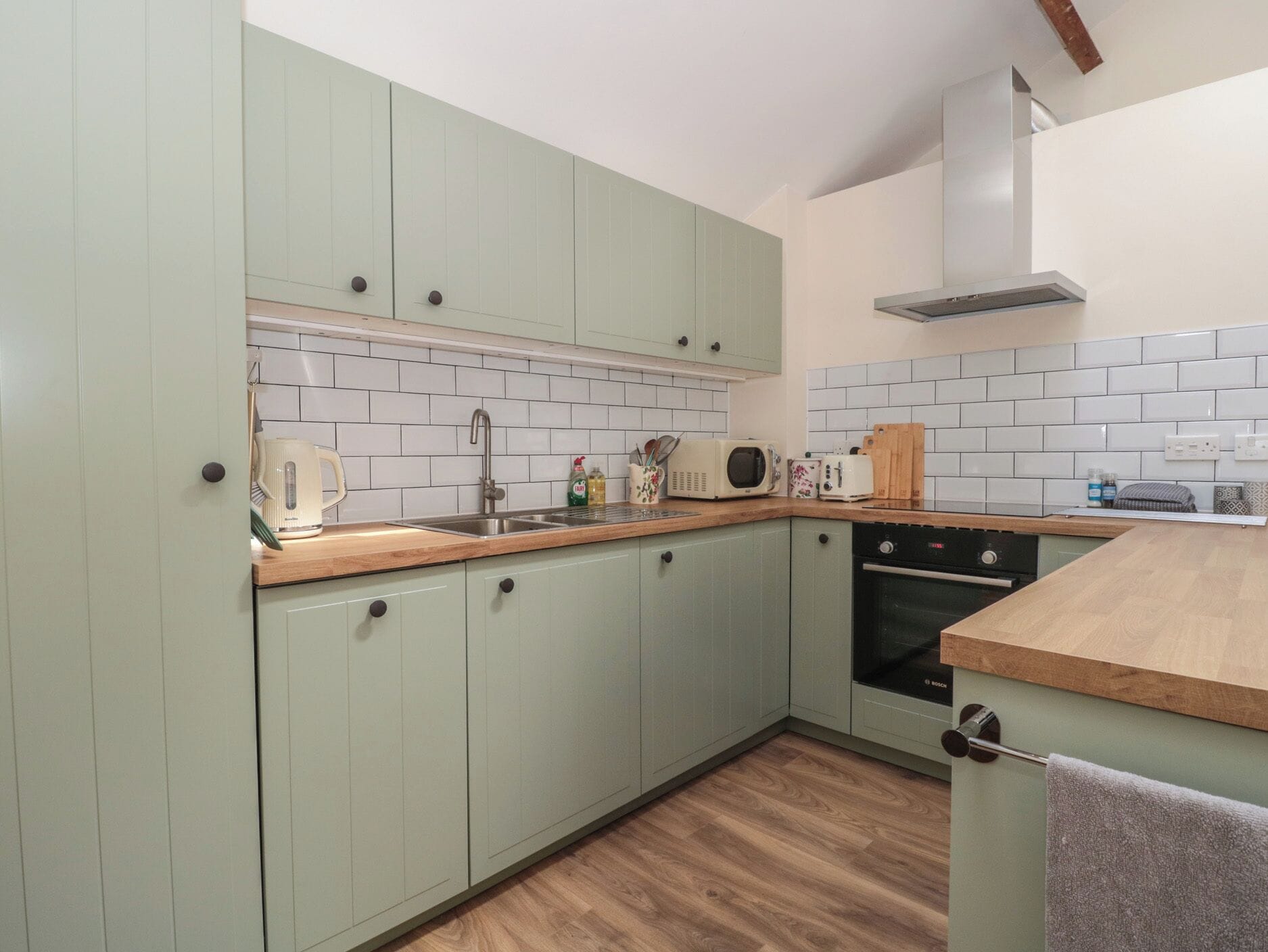 A kitchen with light green cabinets, a wooden countertop, a sink, a stove, and a microwave.