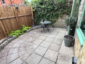 A patio with stone tiles, a wooden fence, a table with chairs, and potted plants. - Wheelwright Cottage (Moreton-in-Marsh)