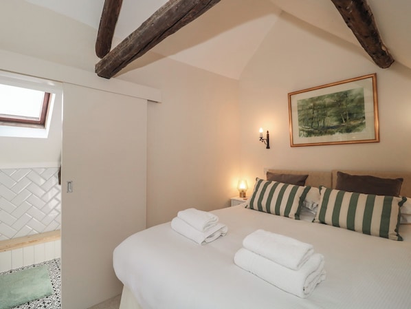 A bedroom with a bed, pillows, a nightstand, a wall-mounted lamp, and a framed picture. - Wheelwright Cottage (Moreton-in-Marsh)