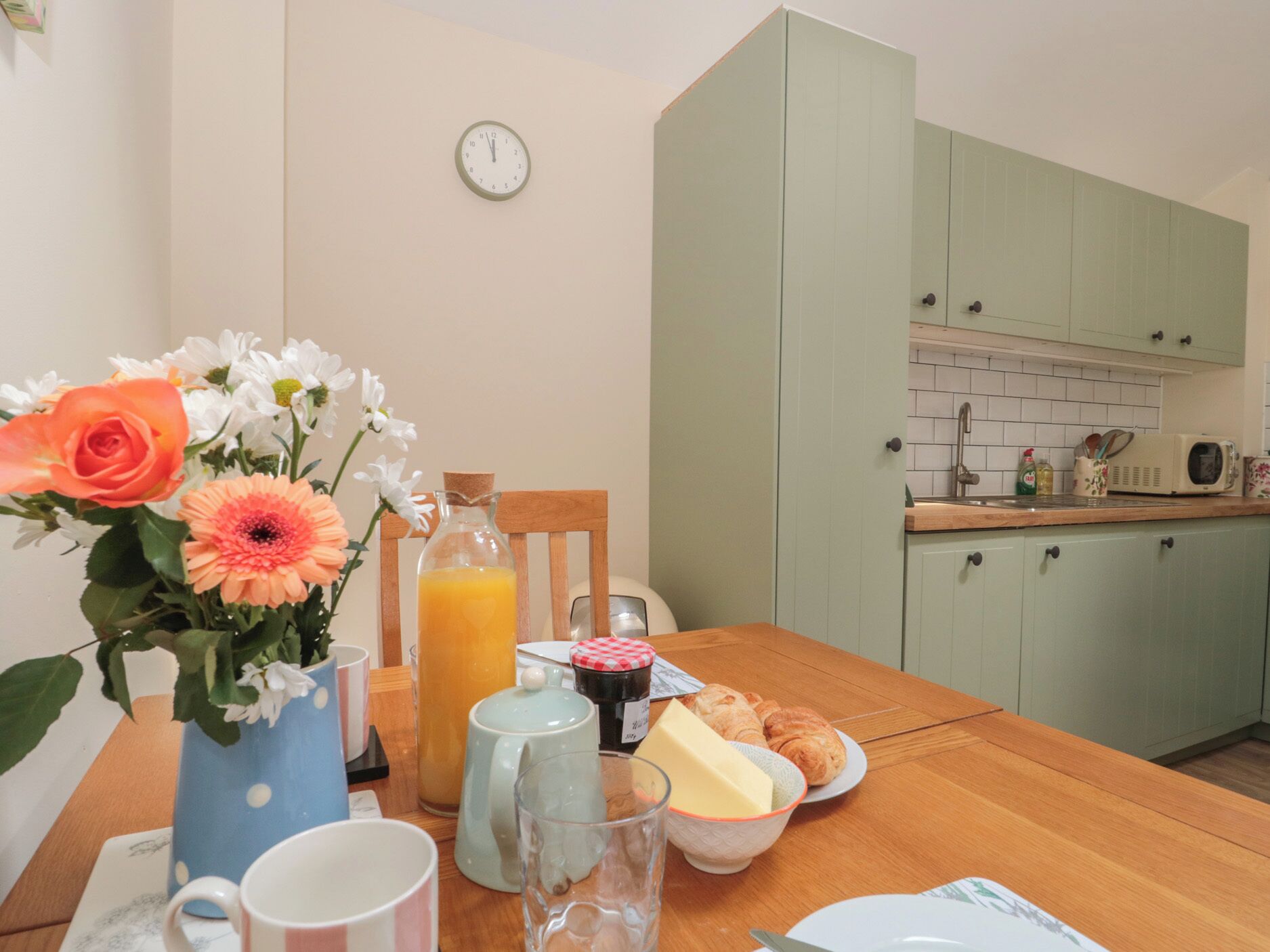 Una cocina con una mesa de madera preparada para el desayuno, incluyendo un jarrón con flores, un jarro de zumo de naranja y un plato con cruasanes.