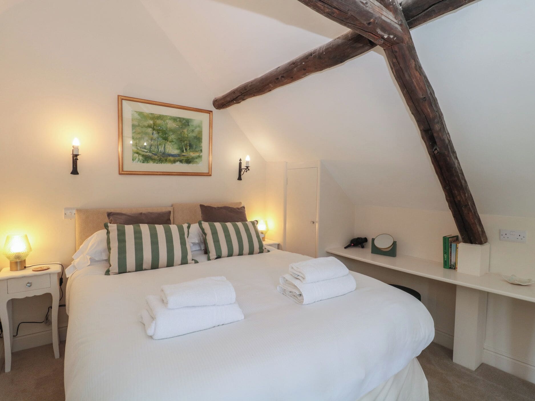 A bedroom with a bed, pillows, a nightstand, a wall-mounted lamp, a framed painting, and a wooden beam.