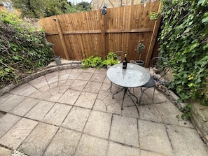 A patio with a round table and chairs, a bottle of wine, and a wooden fence. - Wheelwright Cottage (Moreton-in-Marsh)