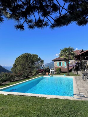 Una piscina con un cielo azul despejado, rodeada de vegetación y un edificio de piedra con techo de tejas.