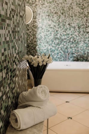 Une salle de bain avec un mur carrelé, une baignoire, un porte-serviettes avec des serviettes pliées et un vase avec des fleurs blanches.