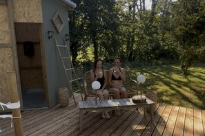 Two people sitting on a wooden deck with a table and chairs, a ladder, and a small building with a door and wall-mounted lights. - Estate with a lake and recreational activities for the whole family: Vacation Rental, Manor House, and Tipis (Montigny-le-Chartif)