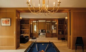 Une table de billard avec des queues de billard, un lustre et des boiseries dans une pièce donnant sur un salon.