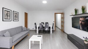 A modern living room with a grey sofa, a white coffee table, a dining area with chairs, a TV mounted on the wall, and a wooden door. - DALI Apartment - Right in the city centre (Salou)