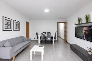 Una sala de estar moderna con un sofá gris, una mesa de centro blanca, un área de comedor con sillas, un televisor montado en la pared y una puerta de madera.