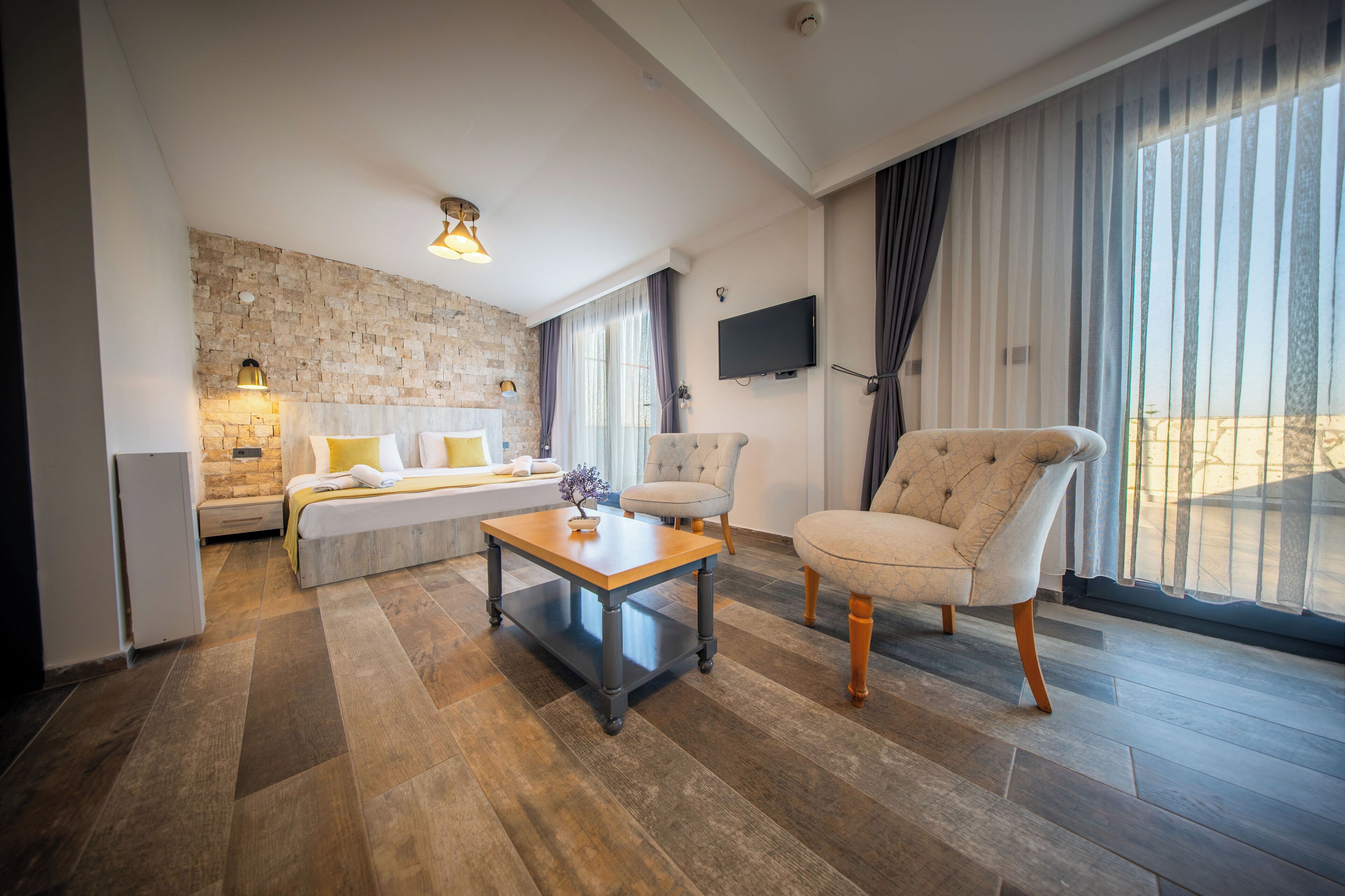 Una habitación de hotel moderna con una cama, una mesita de centro de madera, dos sillones, una pared decorativa de piedra, un televisor de pantalla plana y ventanas grandes con cortinas.