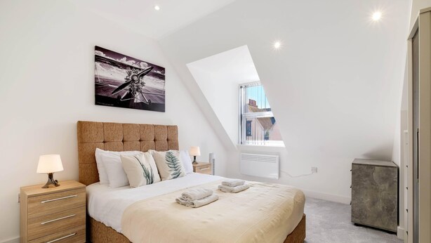 Ein Schlafzimmer mit einem großen Bett, einem Nachttisch mit Lampe, einem Kopfteil, einem Bild an der Wand und einem Dachfenster.