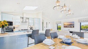 A modern kitchen with a central island, a dining area with a table set for six, and a large window with a view. - Swanlake Lodge - 3 Bedroom Lodge - Penally (Tenby)