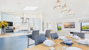 A modern kitchen with a central island, a dining area with a table set for four, and a view of the outdoors through large windows.