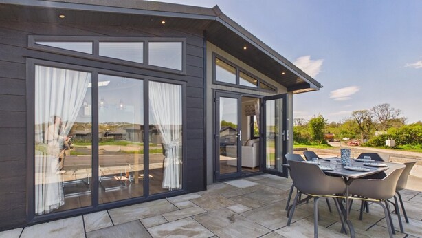 A modern house with large glass doors, a patio with a table and chairs, and a view of the surrounding landscape. - Swanlake Lodge - 3 Bedroom Lodge - Penally (Tenby)