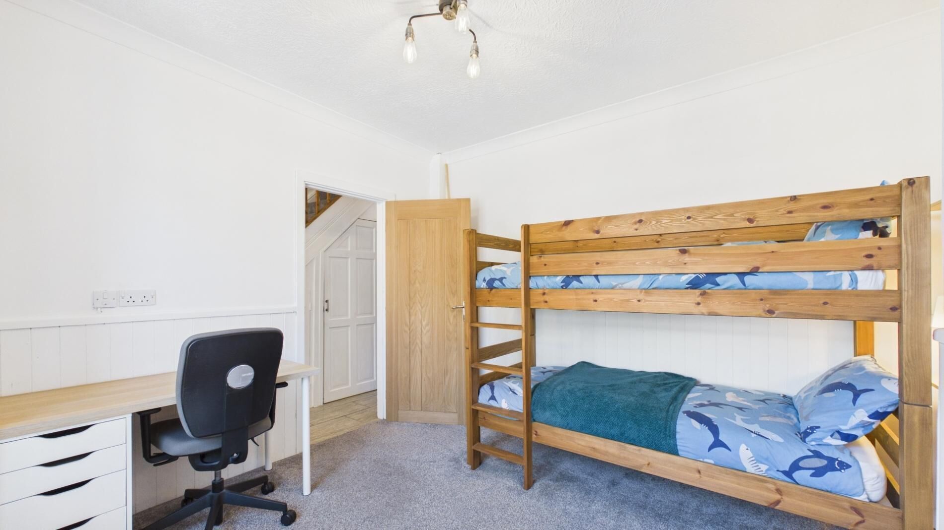 A bunk bed with a desk and chair, a wooden wardrobe, and a door leading to another room.