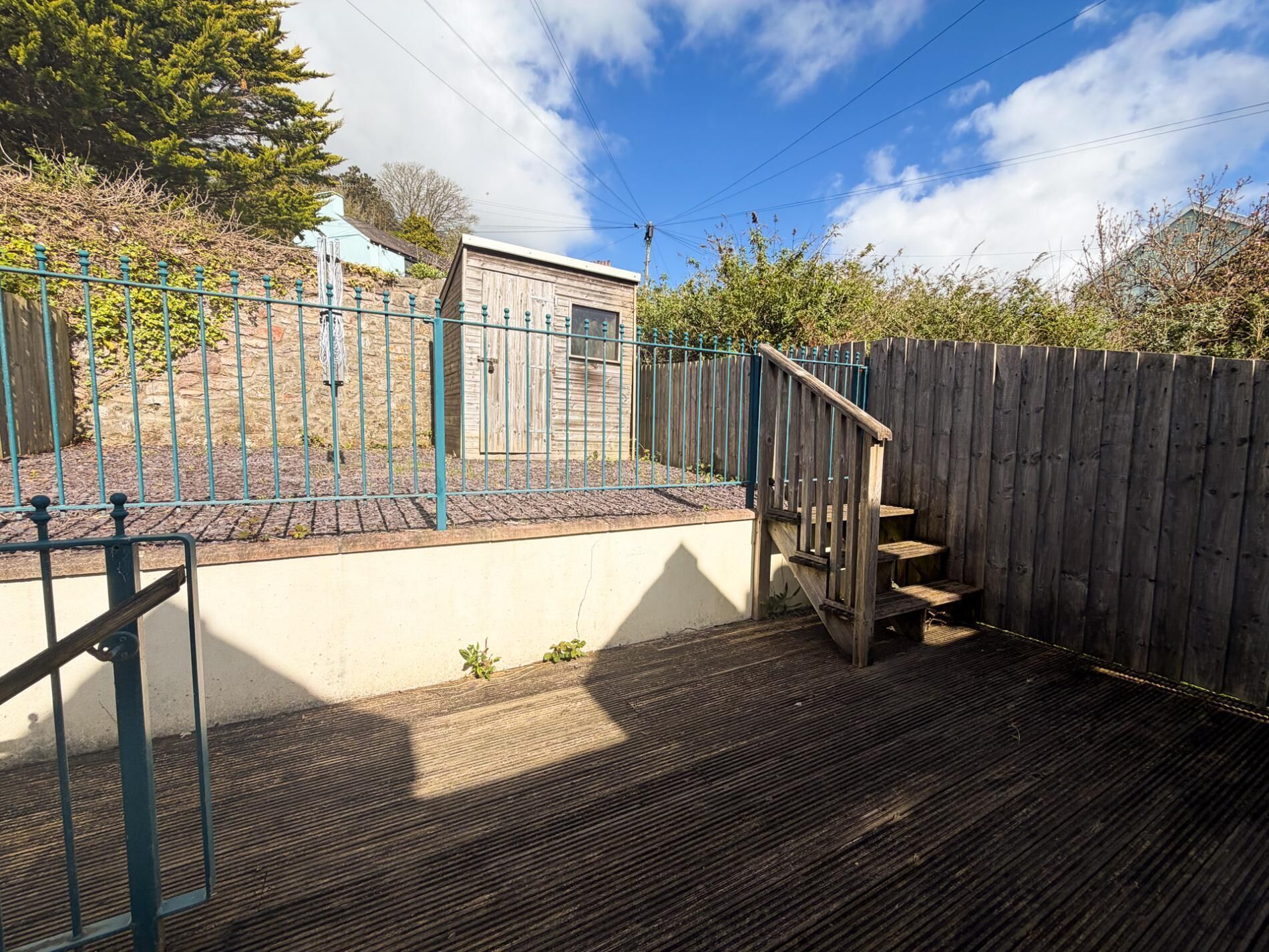 Eine Holzterrasse mit Metallgeländer, ein Holzzaun und ein kleines Gebäude im Hintergrund.