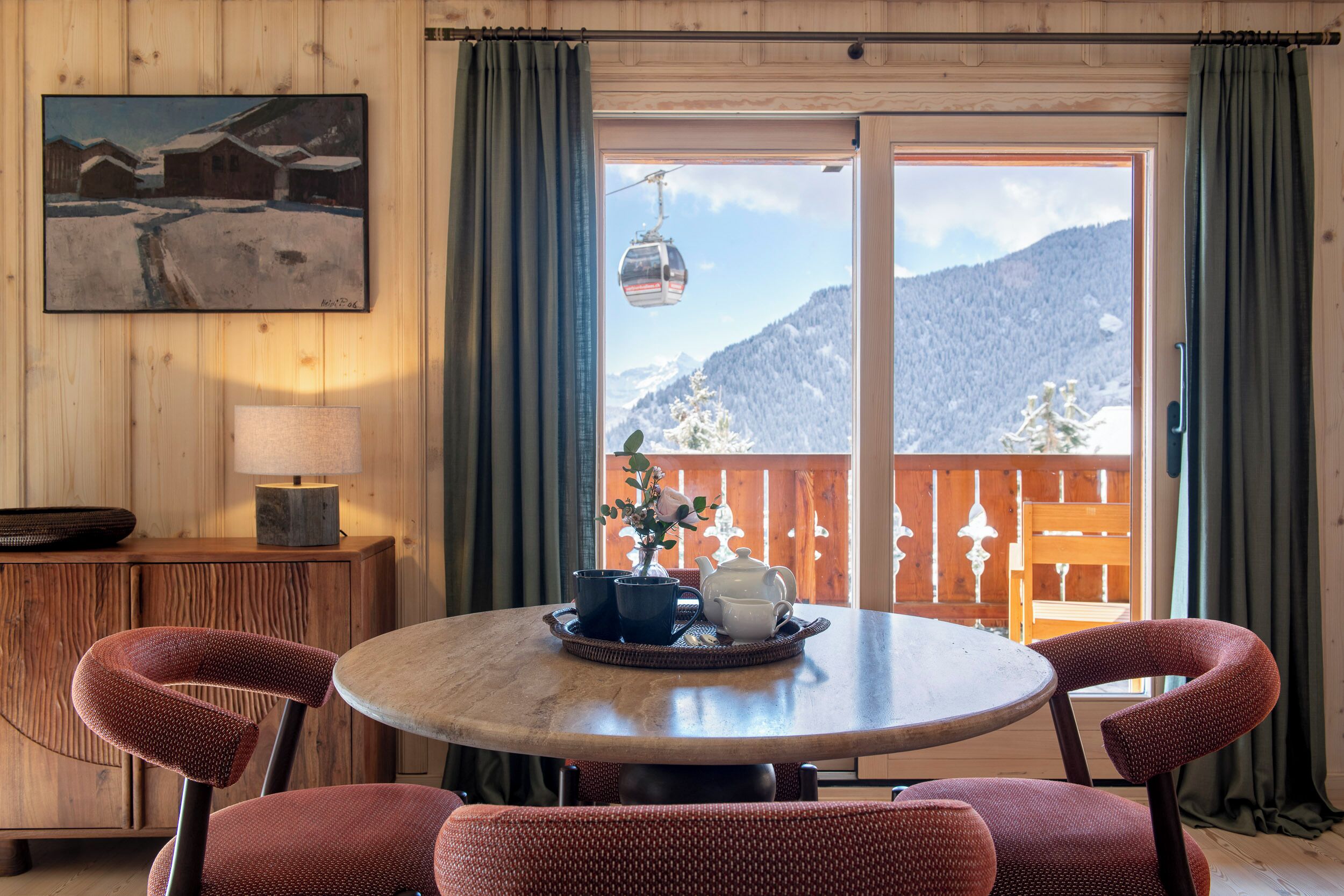 A dining area with a round table, chairs, a painting, and a view of a mountain landscape.