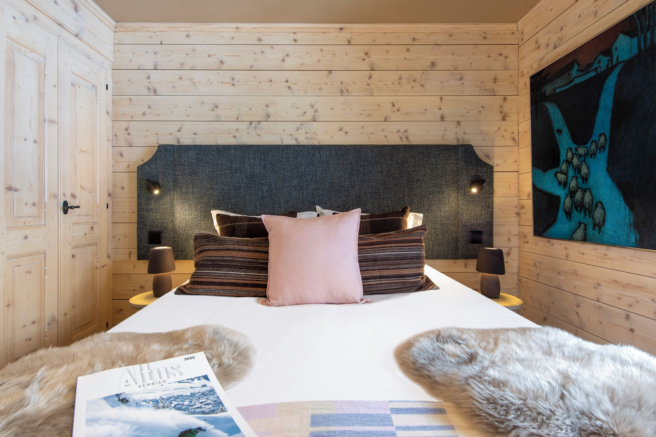 A bedroom with a wooden headboard, a bed with pillows, two bedside lamps, a magazine on the bed, and a painting on the wall.