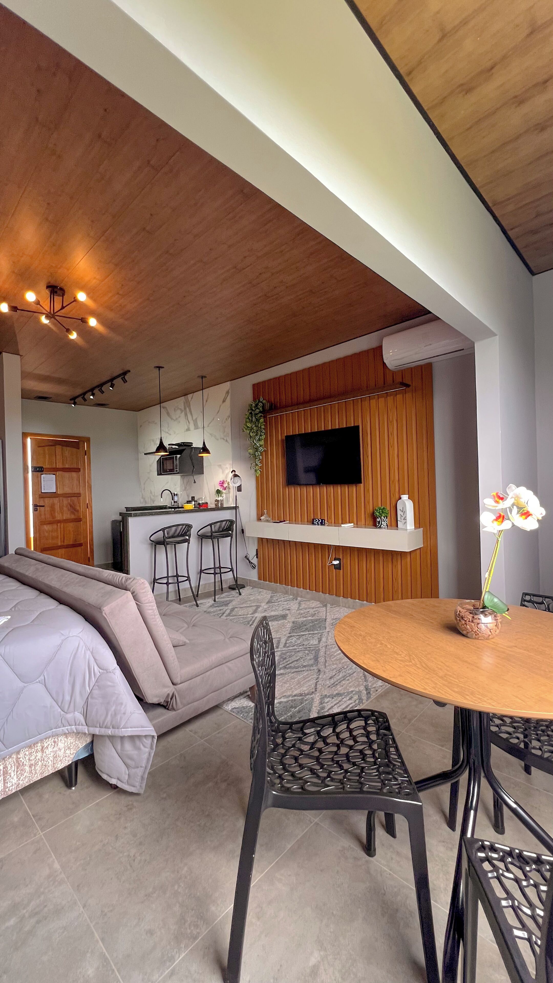 Una sala de estar moderna con sofá, una mesa de comedor con sillas, una zona de cocina con televisor y techo de madera.
