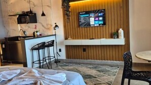 A modern bedroom with a bed, a small kitchenette, a TV mounted on a wooden wall, and a dining area with a table and chairs. - Mirante Vista Nobre (Maricá)
