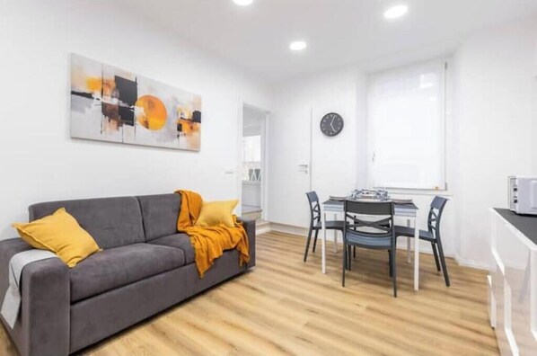 A modern living room with a grey sofa, a dining table with chairs, a clock on the wall, and a large abstract painting.