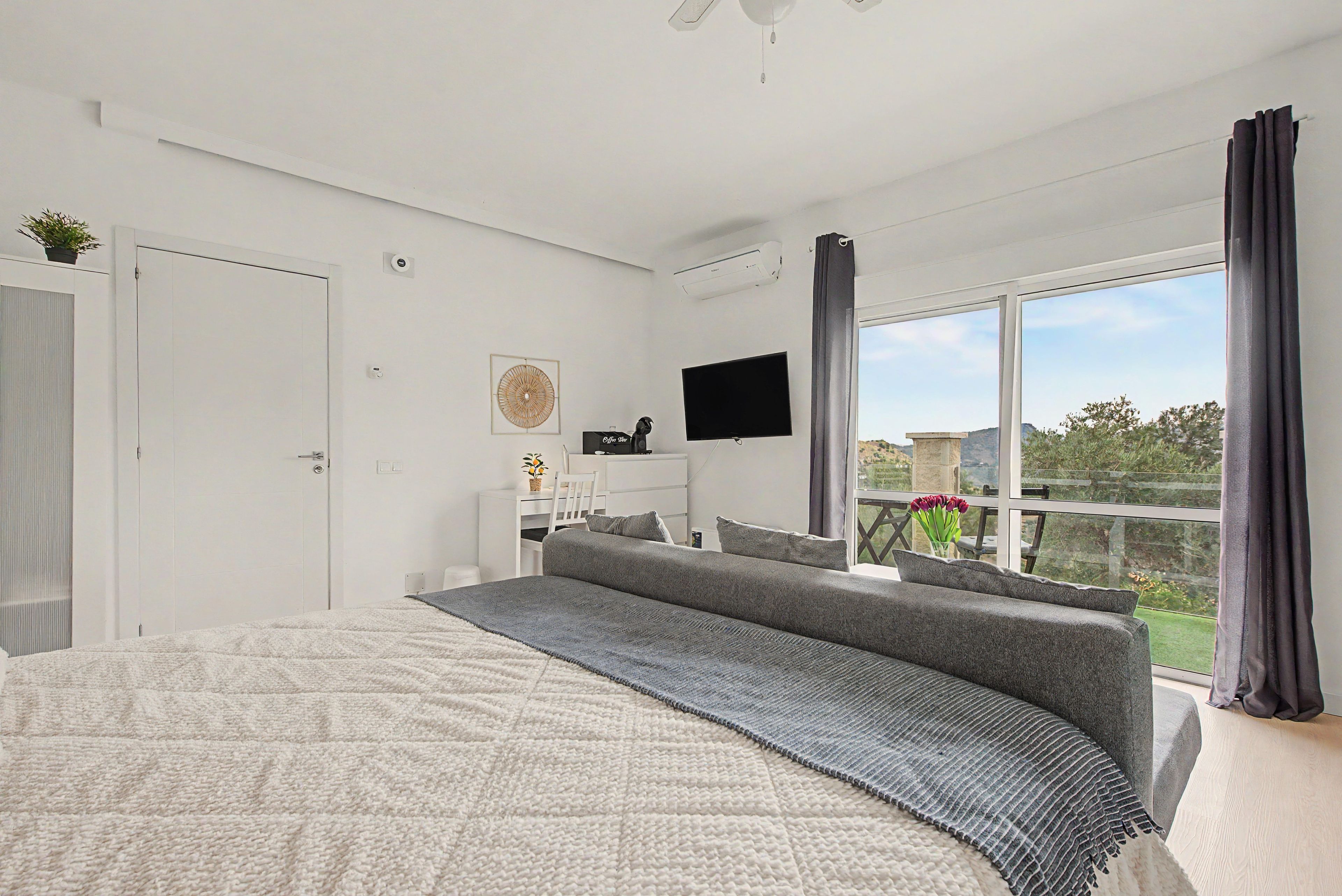 A modern bedroom with a large bed, a flat-screen TV, a desk, and a balcony with a view.
