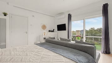 A modern bedroom with a large bed, a flat-screen TV, a desk, and a balcony with a view.