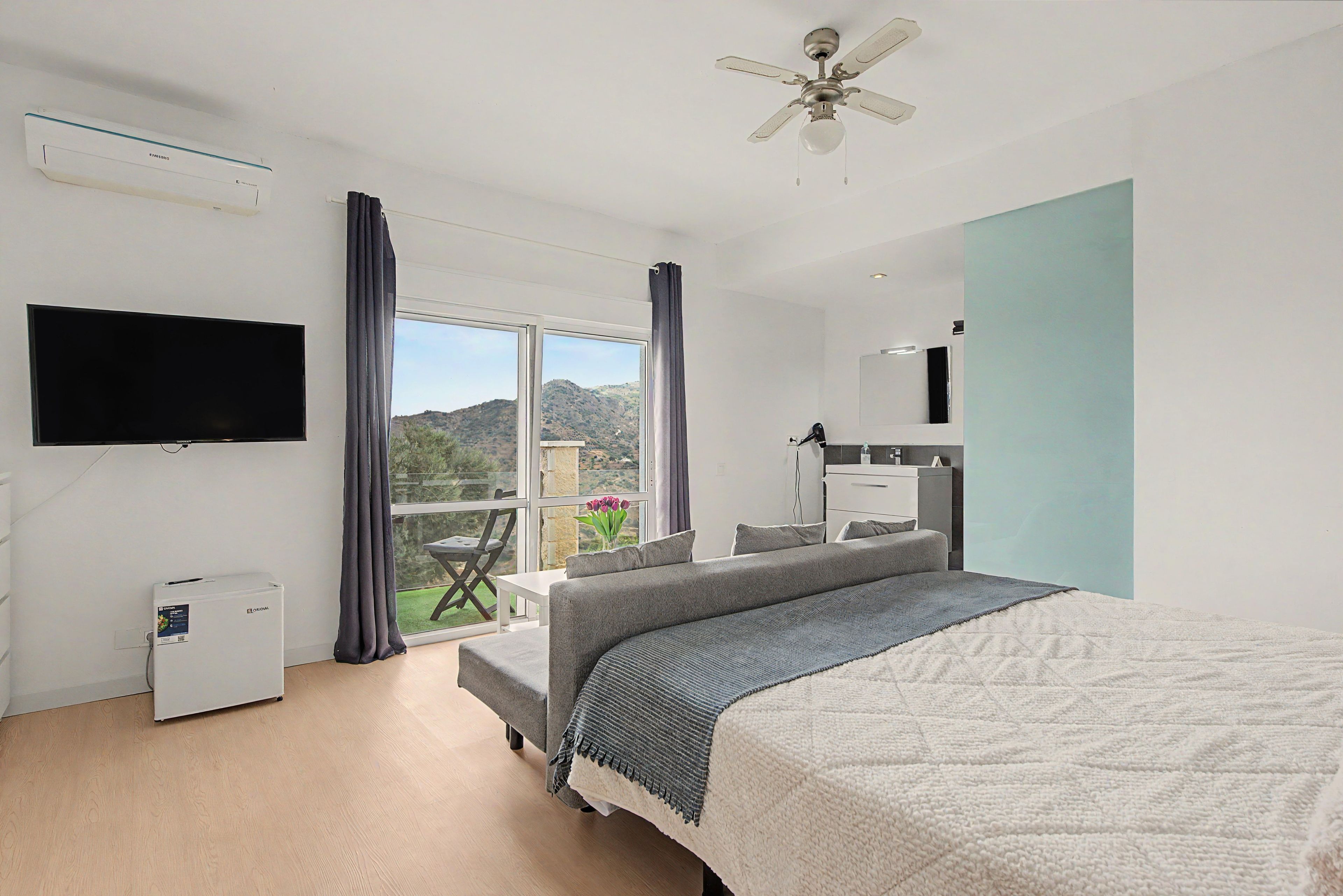A hotel room with a bed, a TV, a ceiling fan, a desk, and a view of the outdoors.
