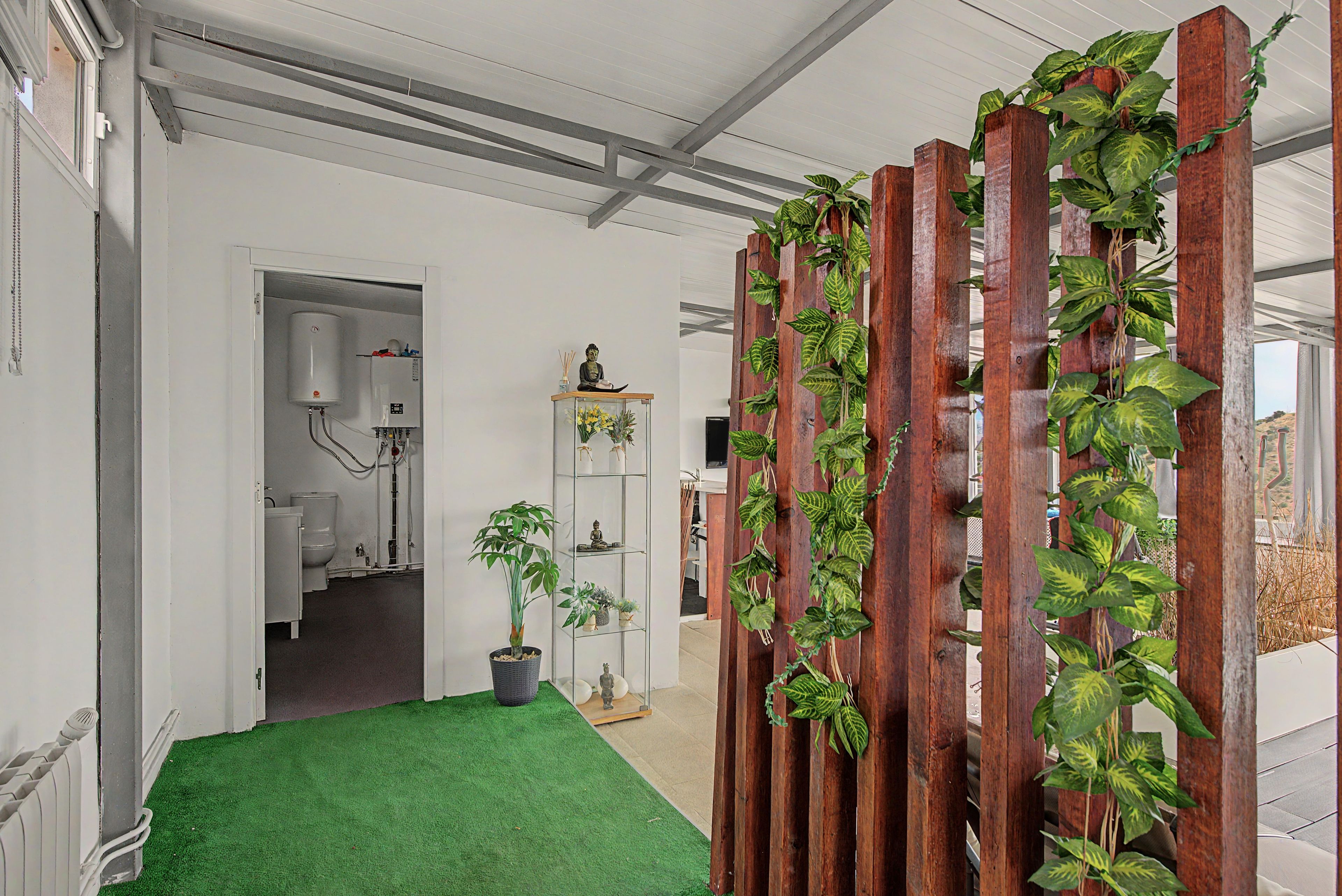 A hallway with a green carpet, a wooden partition with plants, a white bathroom with a toilet, and a shelf with decorative items.