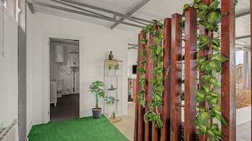A hallway with a green carpet, a wooden partition with plants, a white bathroom with a toilet, and a shelf with decorative items.