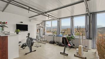 Un gimnasio con equipo de ejercicio, un televisor y vistas a un paisaje montañoso a través de amplias ventanas.