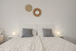Una cama bien hecha con un edredón blanco acolchado, dos almohadas grises y un cabecero. Hay dos mesitas de noche con lámparas, y dos elementos decorativos en la pared sobre la cama.
