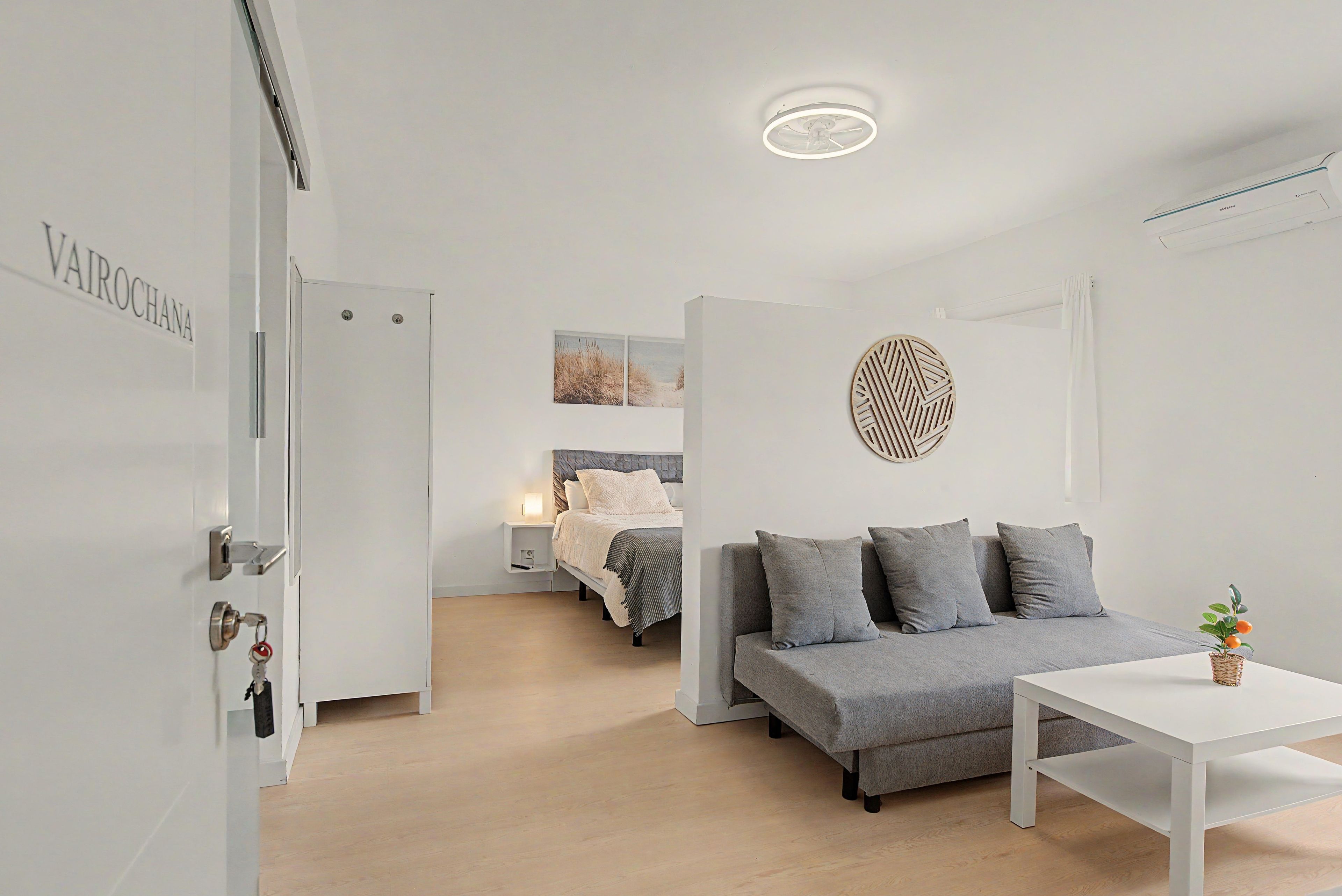 A modern, minimalist hotel room with a grey sofa, a white table, and a bed with a grey headboard.